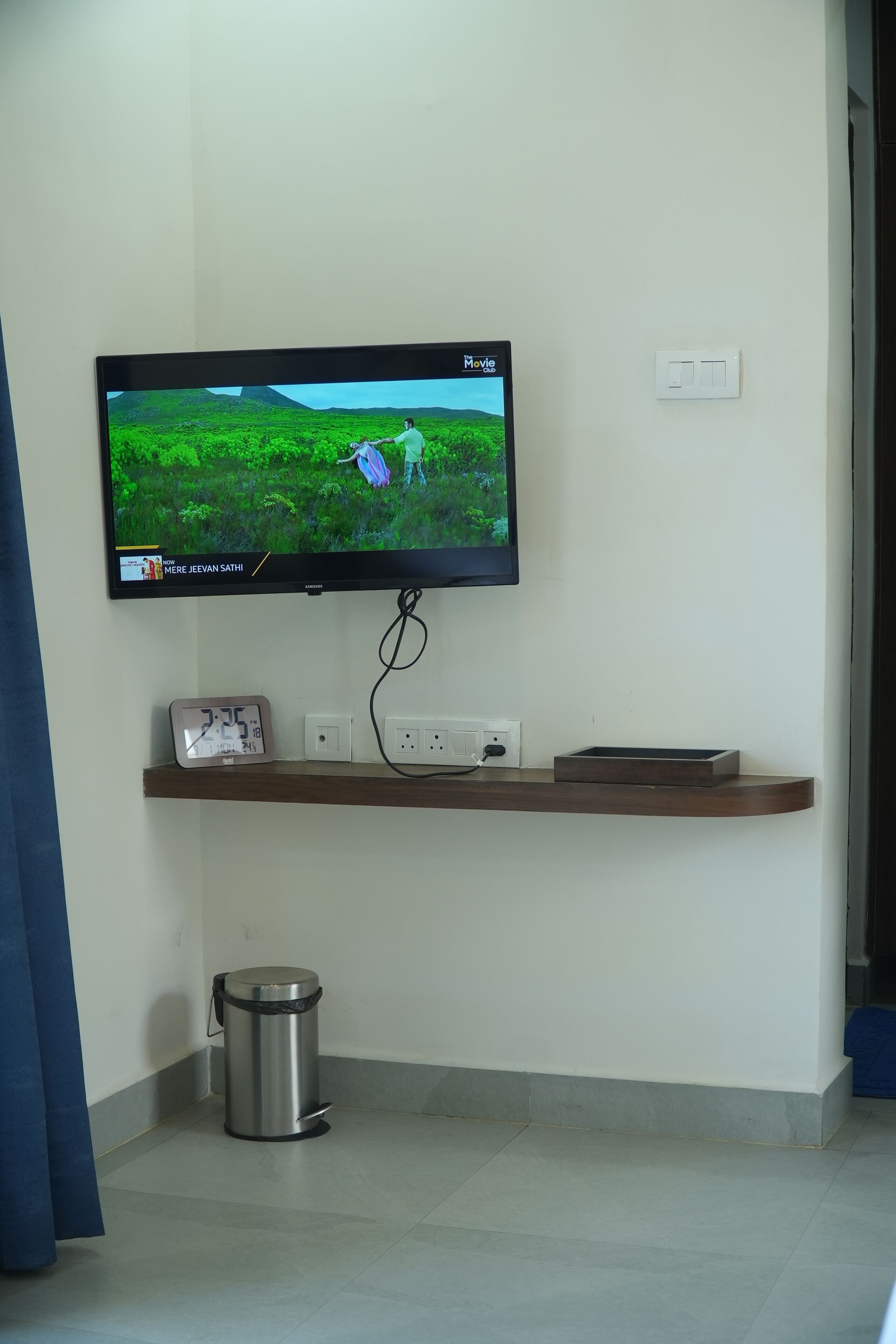 A television mounted on a wall displaying a nature scene. Below the TV, there is a shelf with a clock, a trash can, and a small box.