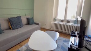 Ett vardagsrum med en soffa, ett vitt soffbord, en tv och ett fönster med utsikt över omgivningen.