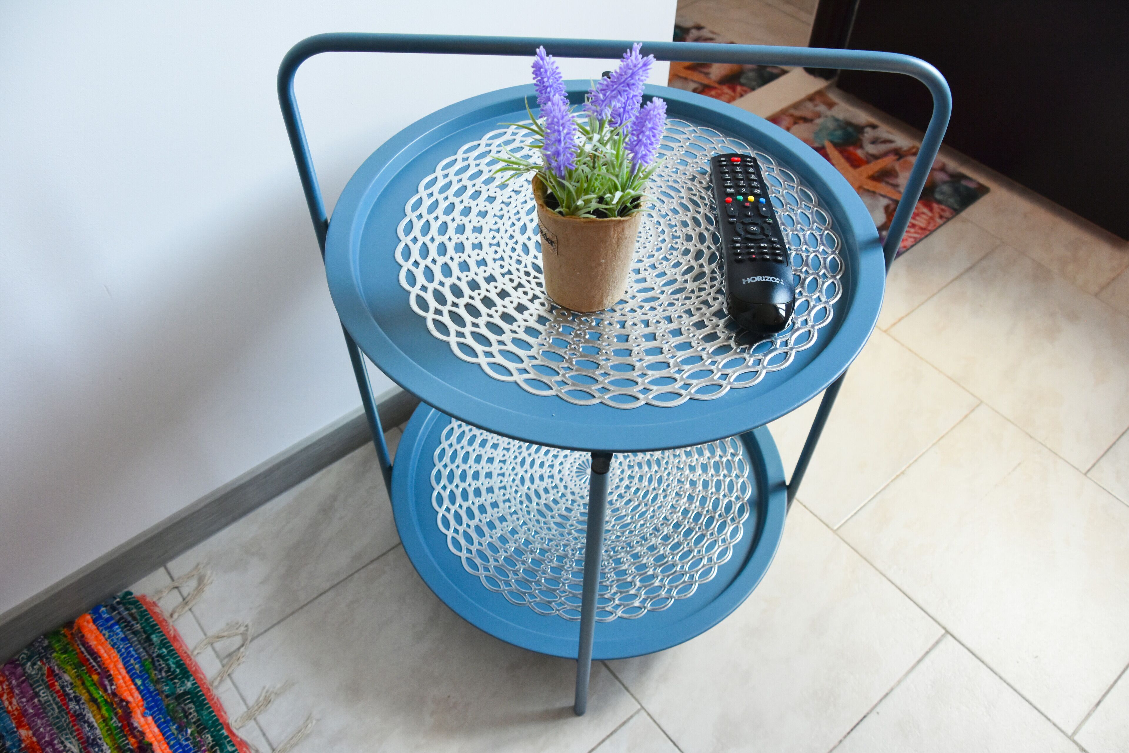 Una mesa metálica azul con superficie trenzada, una planta en maceta con flores moradas y un control remoto.
