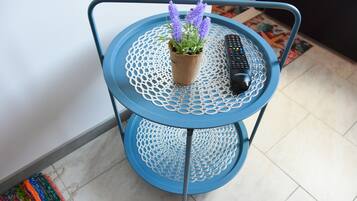 Una mesa metálica azul con superficie trenzada, una planta en maceta con flores moradas y un control remoto.