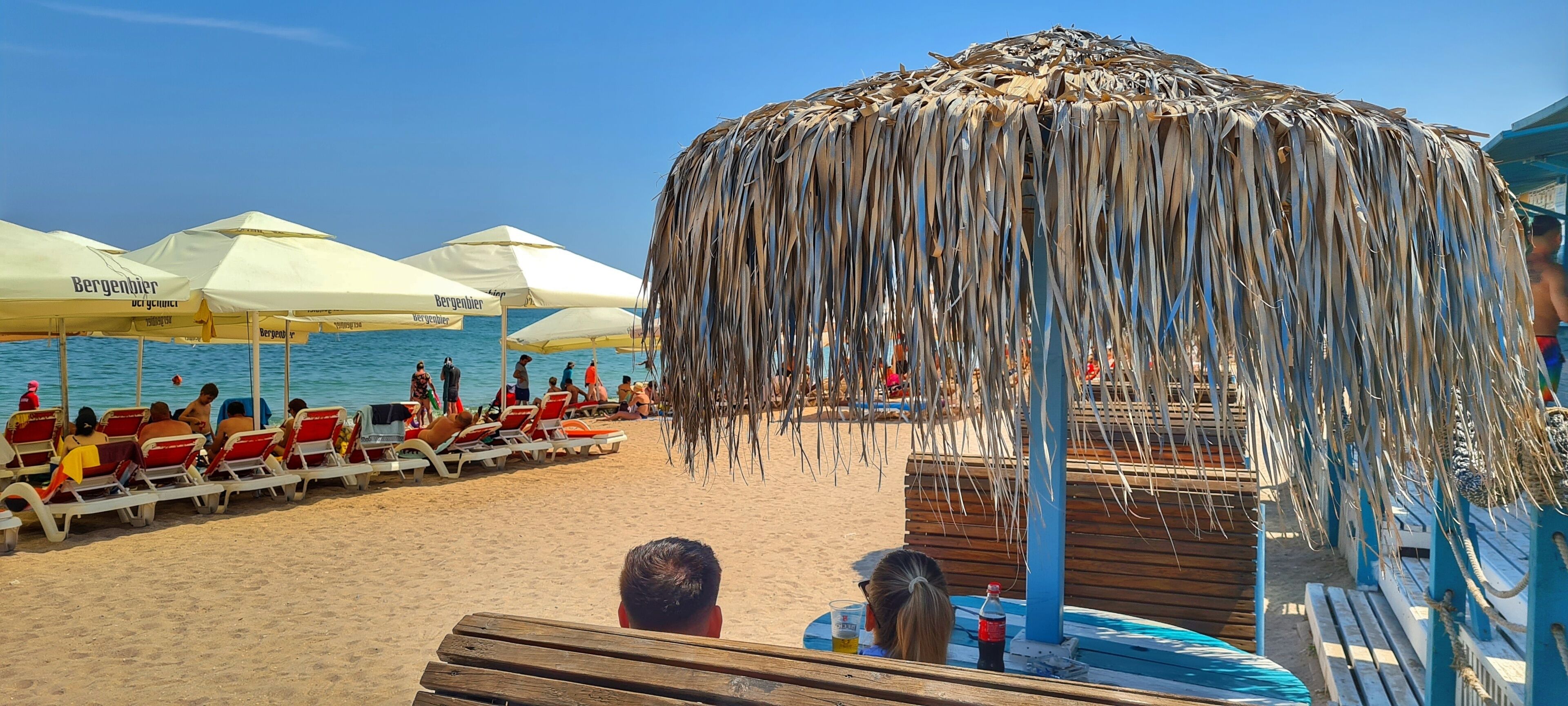 Une plage avec des chaises longues et des parasols, une terrasse en bois et une structure recouverte de chaume.