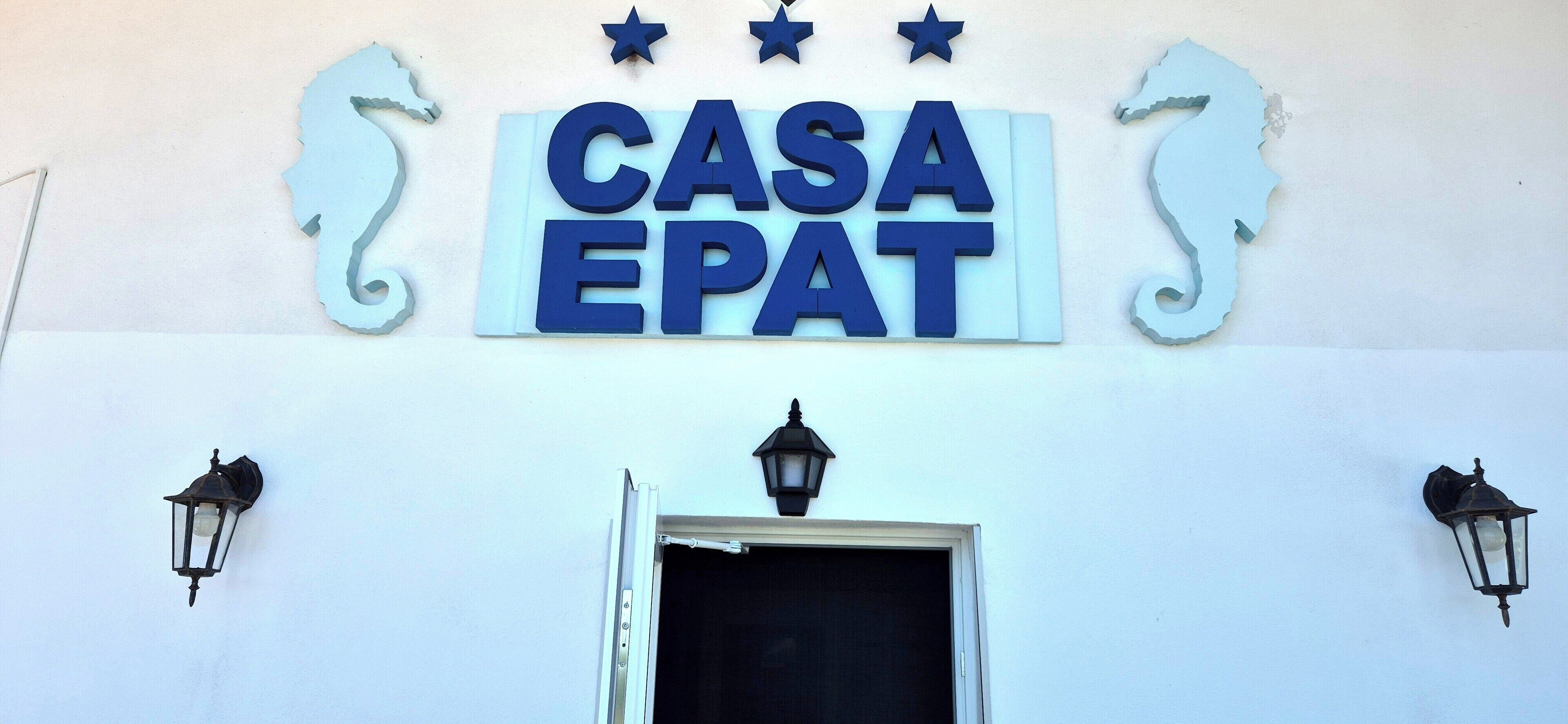 Um letreiro com o texto 'CASA EPAT' acima de uma porta, ladeado por duas lanternas fixadas na parede.