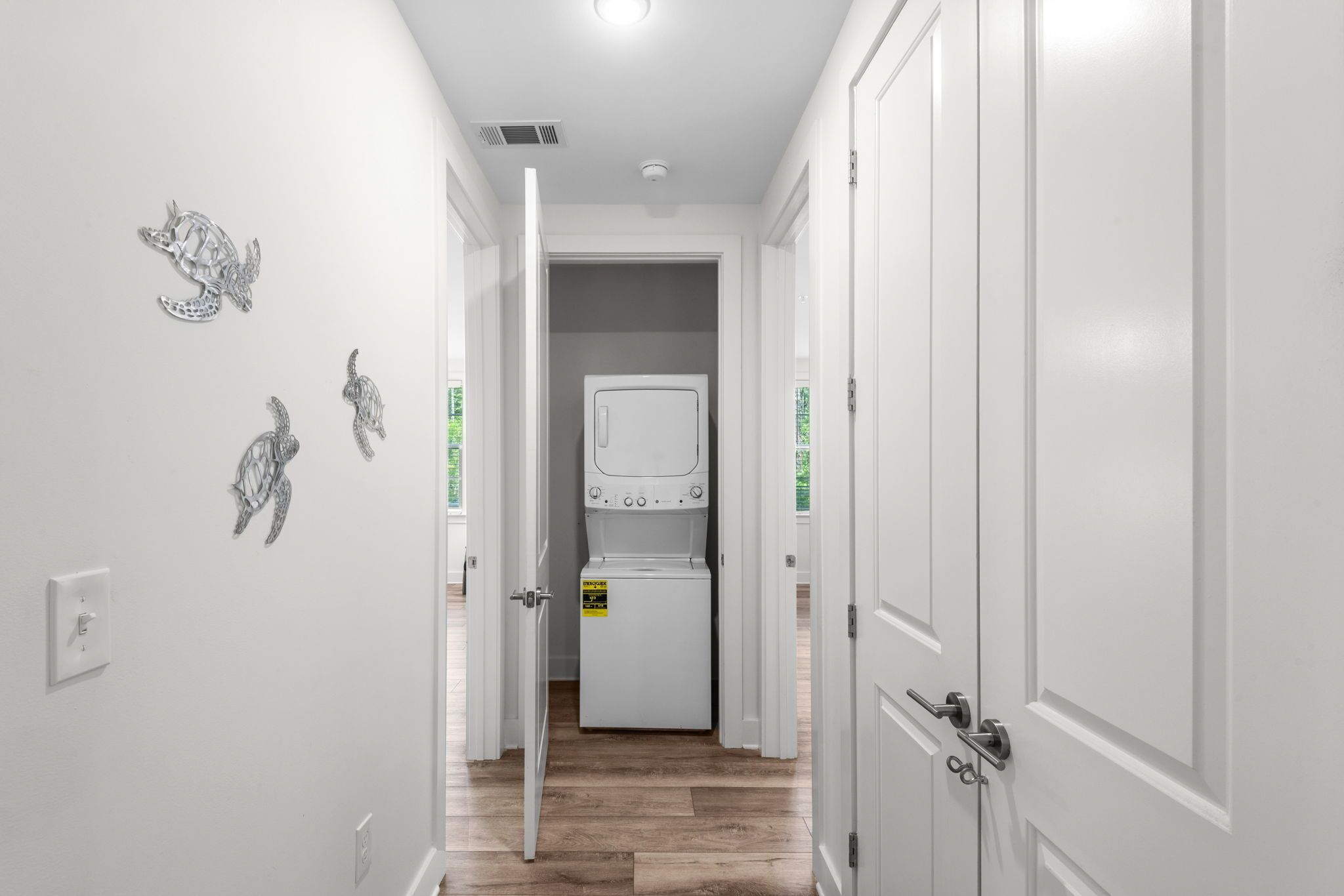 Un couloir aux murs blancs, doté d’un plancher en bois et comprenant une buanderie avec un sèche-linge blanc et des articles empilés.