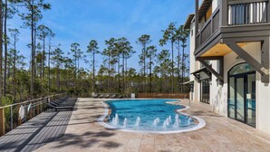 A swimming pool with a fountain feature, surrounded by a wooden deck and a row of lounge chairs. Tall trees are visible in the background. - 2BR | 2BA Close to Beach, Luxury Amenities + Free Tix: Dolphin Cruise & More! (Santa Rosa Beach)