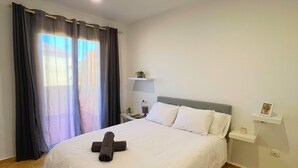 Un lit bien fait, avec une serviette pliée, une table de chevet avec une lampe et une petite plante, et une fenêtre avec des rideaux.