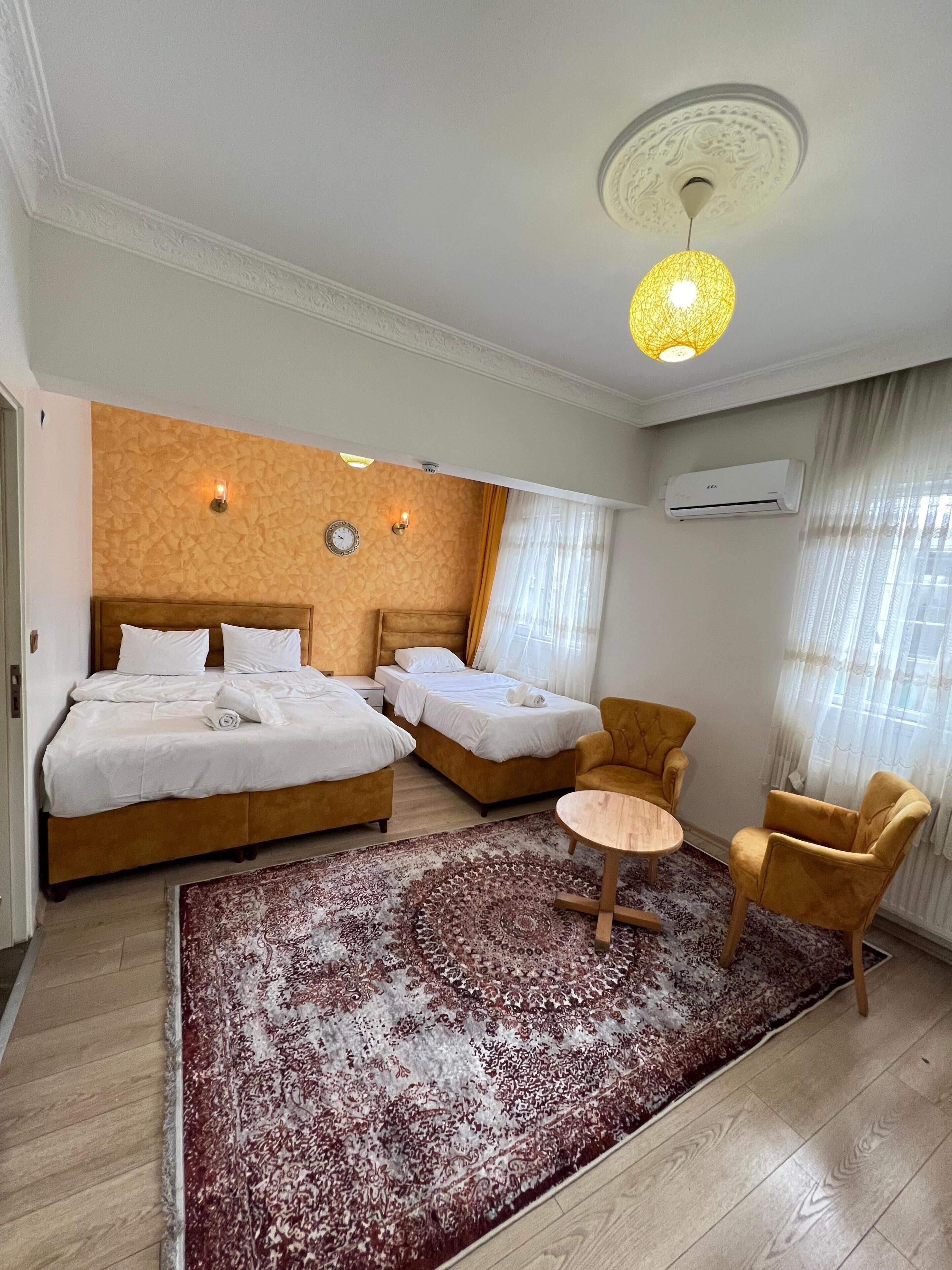 Quarto de hotel com duas camas, uma mesa redonda, duas poltronas, um tapete estampado, um relógio e um aparelho de ar condicionado.