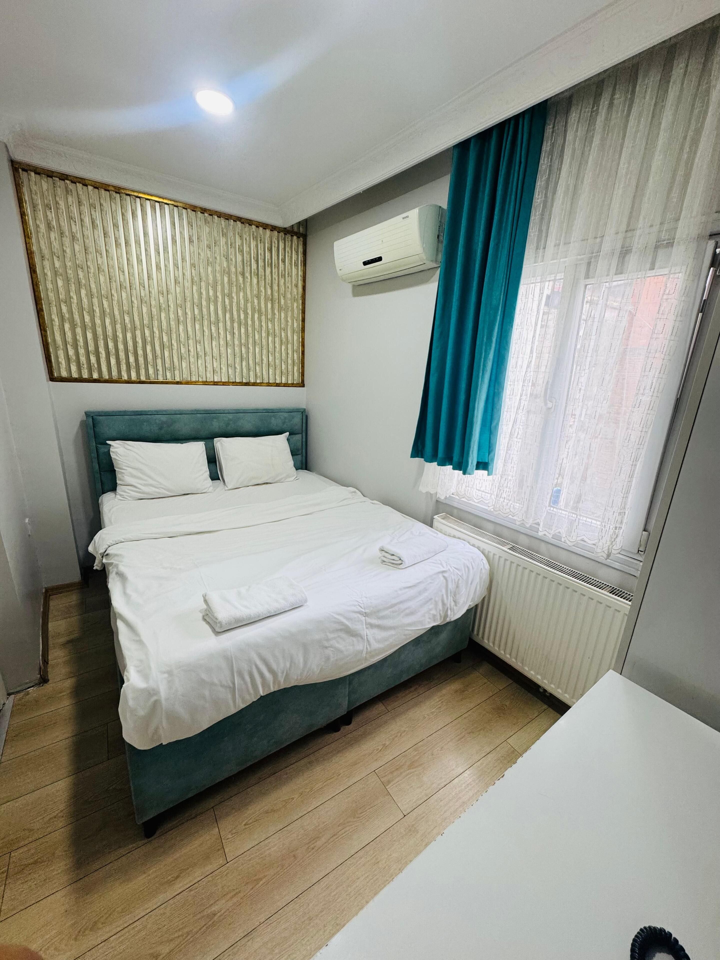 Un lit bien fait, avec du linge de lit blanc, une tête de lit turquoise, une fenêtre avec des rideaux bleus et un radiateur.
