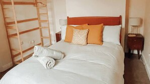 Una cama bien hecha con sábanas blancas y dos almohadas naranjas, una escalera de madera contra la pared, y una mesita de noche con una lámpara.