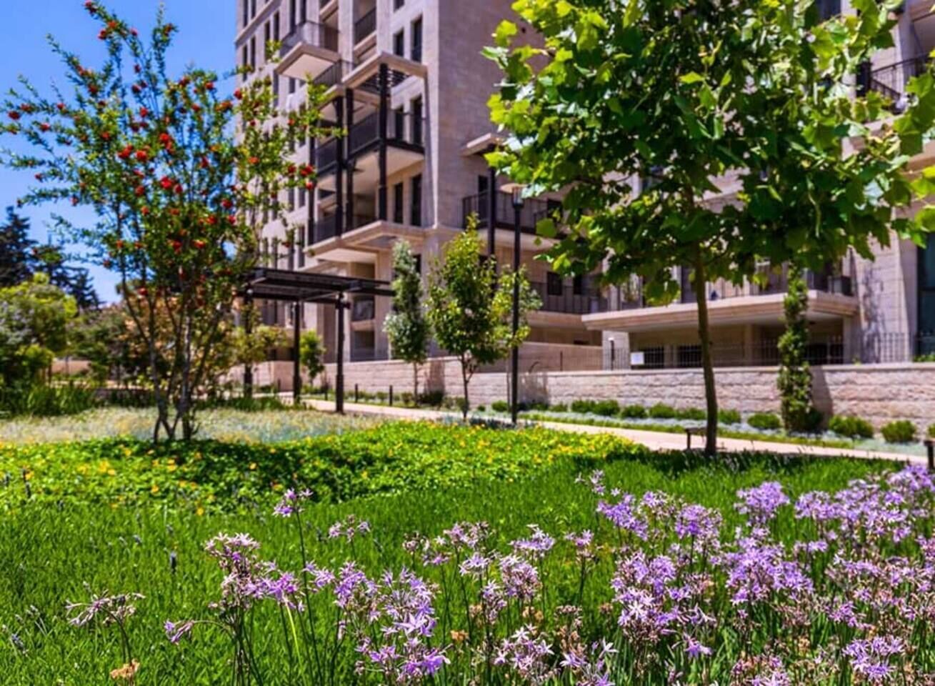 Un bâtiment moderne doté de balcons, entouré d’un jardin bien entretenu où poussent des fleurs violettes.