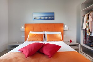 Une chambre d’hôtel comprenant un lit, deux tables de chevet, une commode, une lampe fixée au mur, un tableau et un porte-vêtements.