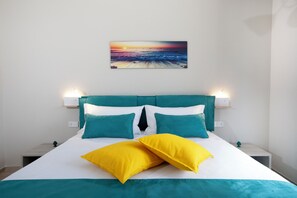 Ein Bett mit türkisfarbener und weißer Bettwäsche, zwei gelbe Kissen und ein Sonnenuntergangsgemälde an der Wand.