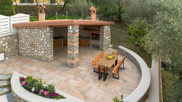 Une terrasse en pierre avec une table et des bancs en bois, entourée d’un muret et d’un espace agrémenté d’un banc incurvé.