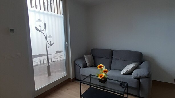 Un salon avec un canapé gris, une table basse ornée d’un vase de tournesols, et une vue sur une balustrade et l’eau, visible à travers une grande fenêtre.
