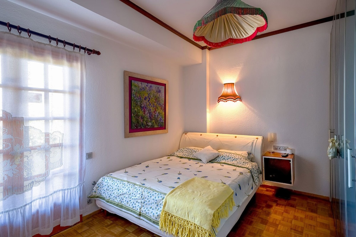 Une chambre à coucher avec un lit, une table de chevet avec une lampe, une fenêtre avec des rideaux et un tableau accroché au mur.