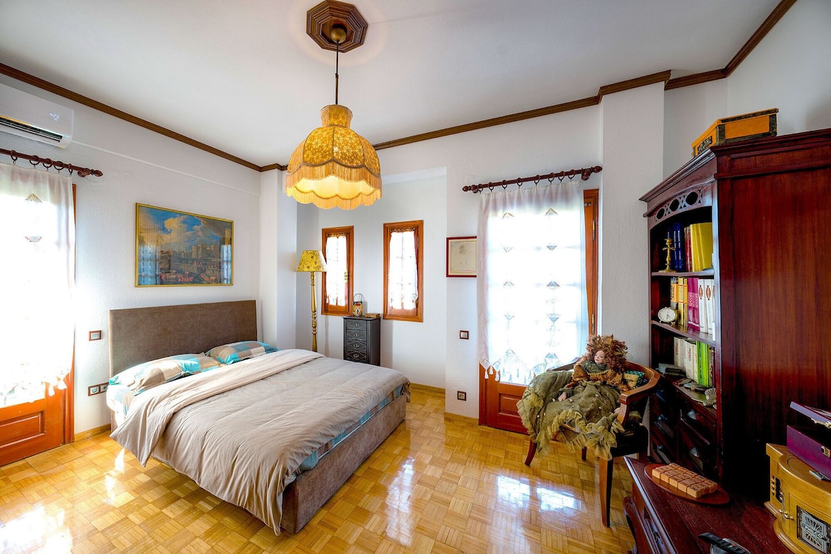 Une chambre avec un grand lit, un plancher en bois, un ventilateur de plafond, une bibliothèque et une fenêtre avec des rideaux.