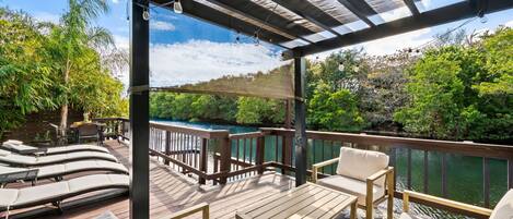 A covered outdoor deck with lounge chairs and a table, overlooking a body of water and lush greenery.