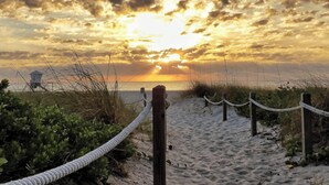 Ein Sandweg am Strand mit Seilspindeln, der zu einem Strandabschnitt führt, im Hintergrund ist ein Rettungsturm zu sehen.