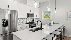 A modern kitchen with white cabinets, stainless steel appliances, a large island with a sink, and pendant lighting. - Pet Friendly Beach House - Bella View (Port St. Joe)