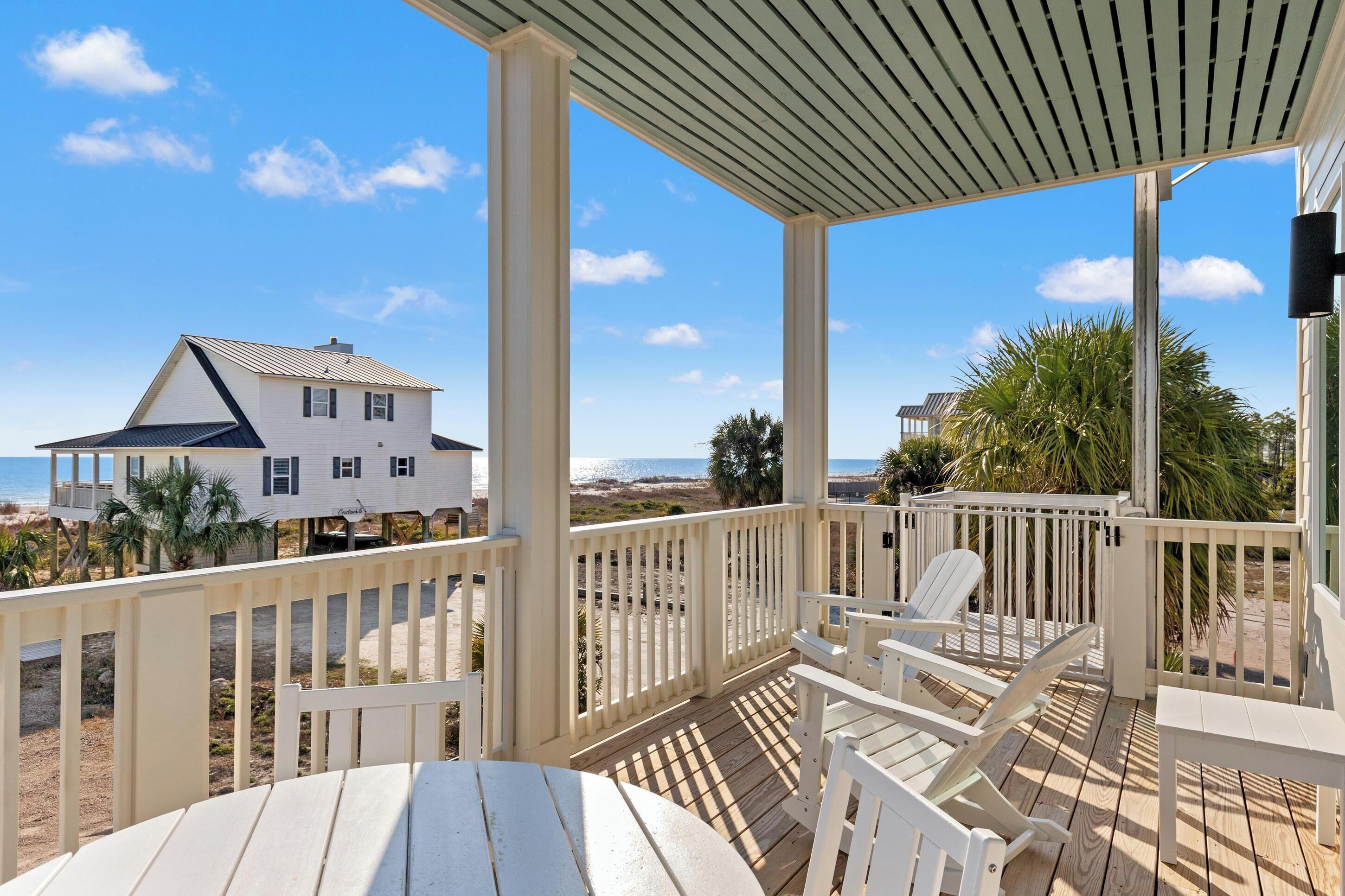 A covered deck with white chairs and a table, overlooking a beach and a white house.