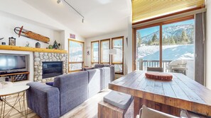 A modern living room with a stone fireplace, a television, a large sofa, and a dining table with a wooden top. - Spacious Renovated Corner Townhome, Laundry, Garage, BBQ (Mammoth Lakes)