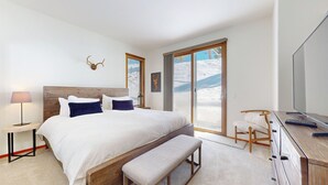 Ein modernes Schlafzimmer mit einem großen Bett, einem Holzkopfteil, einer Bank, einem Schreibtisch mit Fernseher und Blick auf eine verschneite Landschaft durch das Fenster.