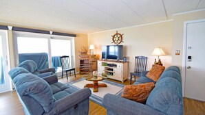 A living room with two sofas, a coffee table, a TV, a bookshelf, and a door. - Thunder Island 13a 2 Bedroom Condo (Ocean City)