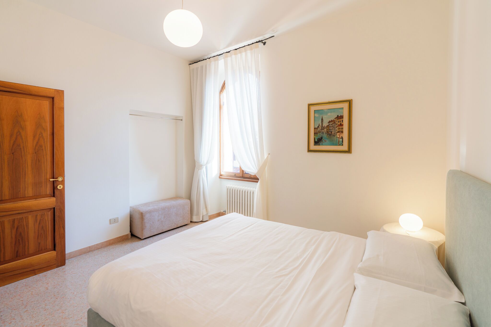 A bedroom with a bed, a nightstand with a lamp, a window with curtains, a radiator, and a door.