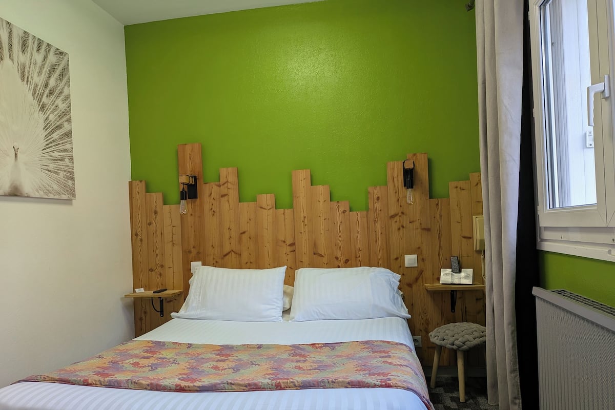 A room with a bed, a green wall, a wooden headboard, a small stool, and a window with curtains.