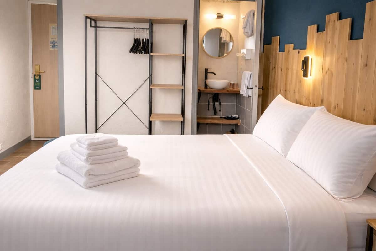 A hotel room with a bed, a wooden wardrobe, a bathroom with a sink and mirror, and a wall-mounted towel rack.
