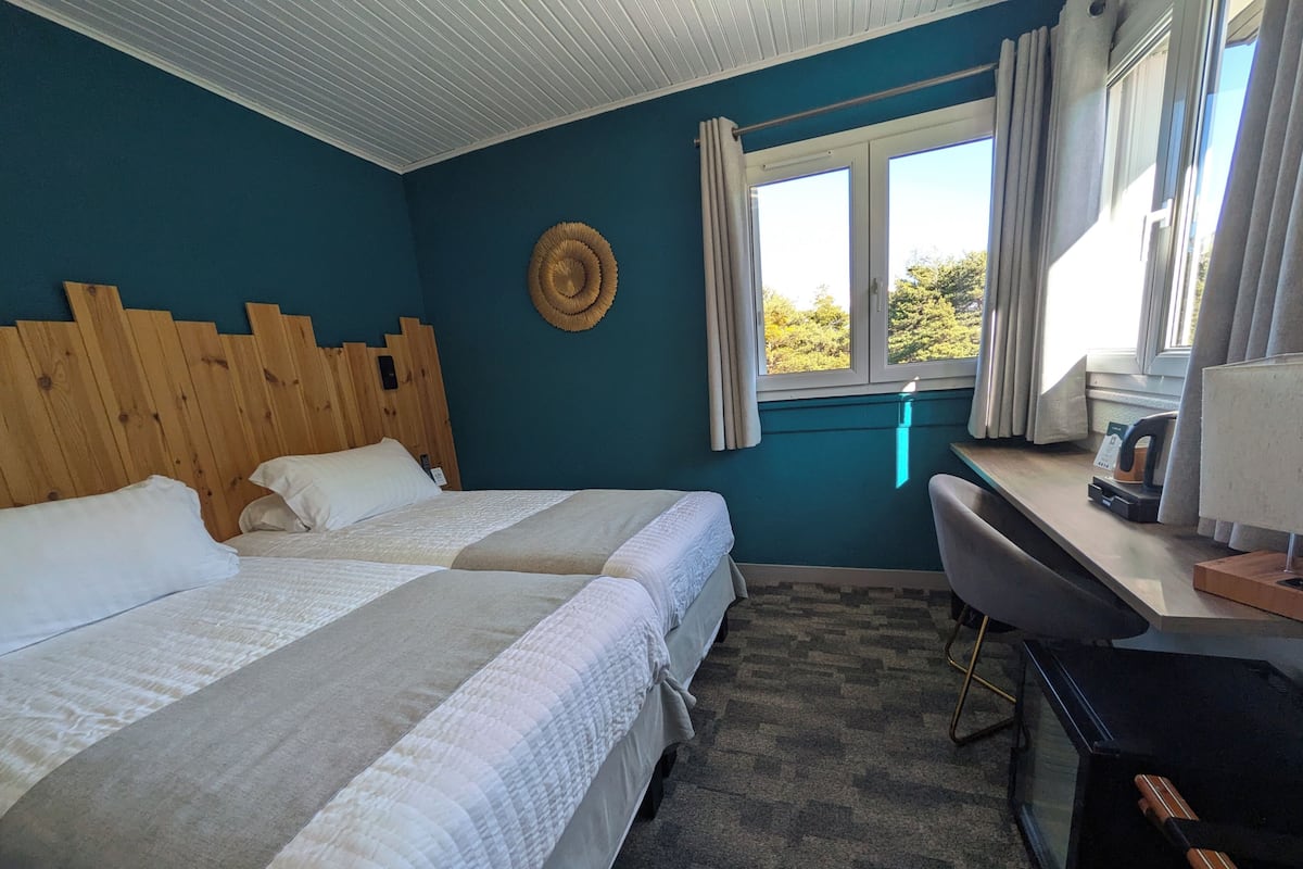 A room with two beds, a desk, a chair, a window with curtains, and a wooden headboard.