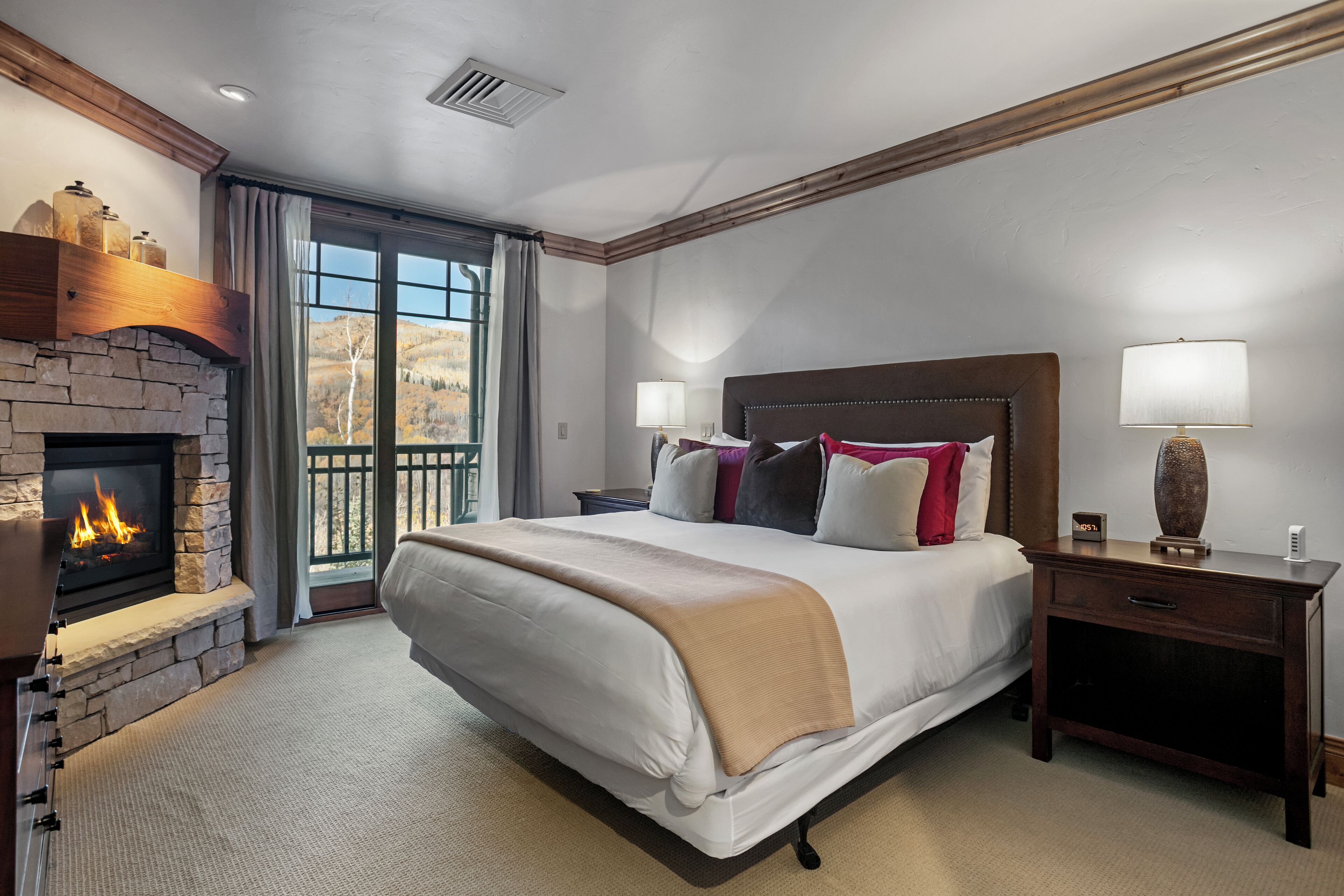 A spacious bedroom with a large bed, a stone fireplace, a nightstand, and a view of the outdoors.