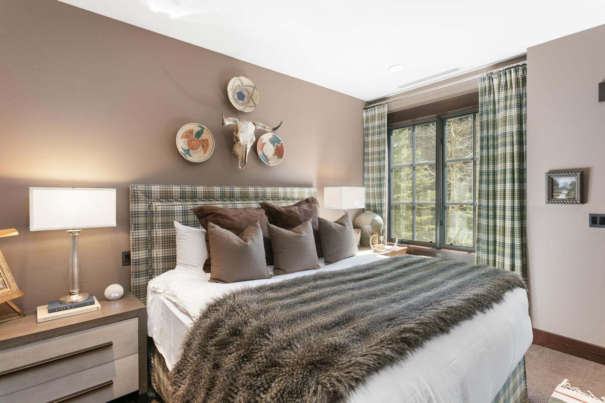 Une chambre avec un grand lit, une table de chevet avec une lampe, une fenêtre avec des rideaux et des éléments de décoration murale, notamment un crâne et des assiettes.