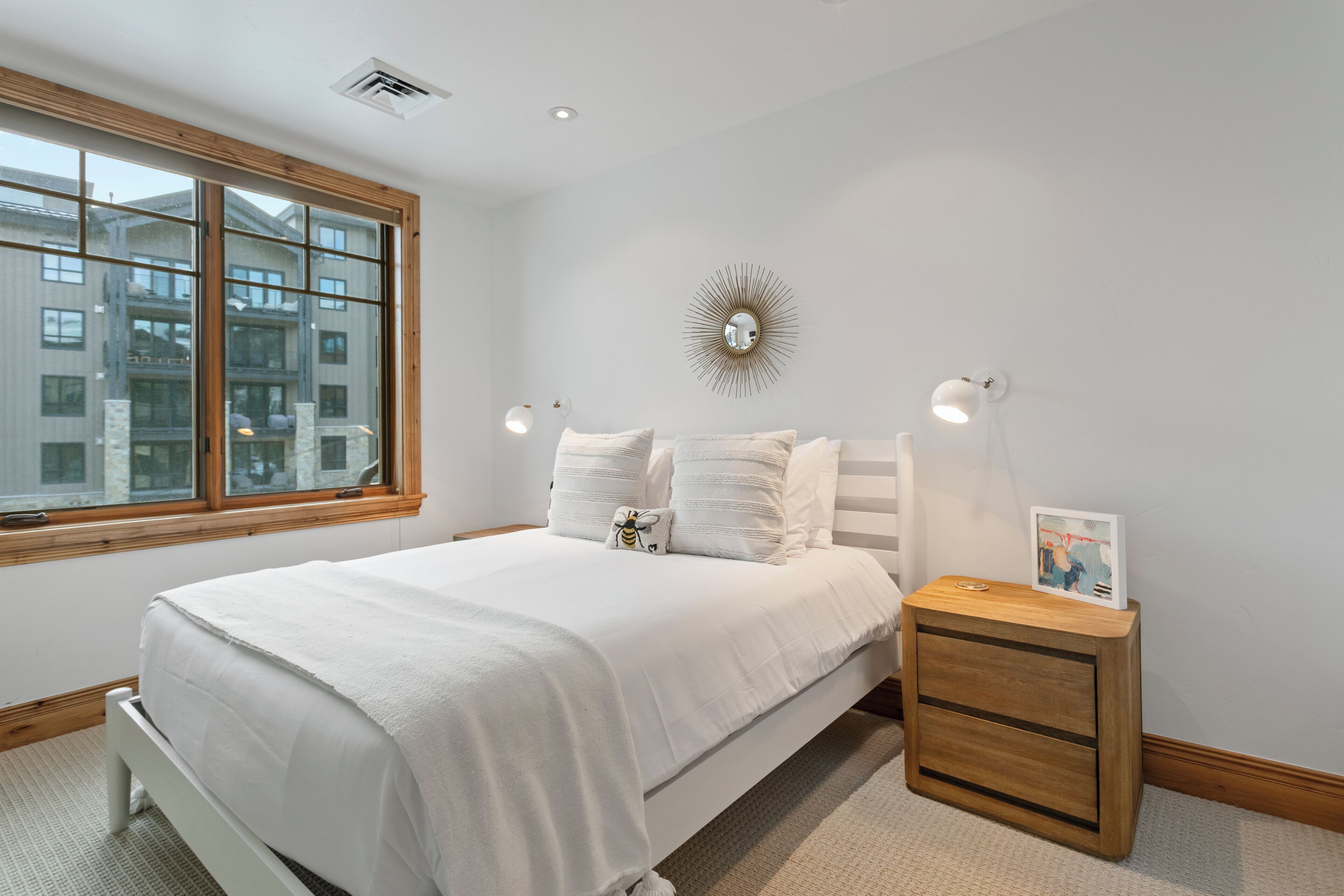 A bedroom with a large window, a bed with white bedding, a bedside table with a lamp, and a wall-mounted mirror.