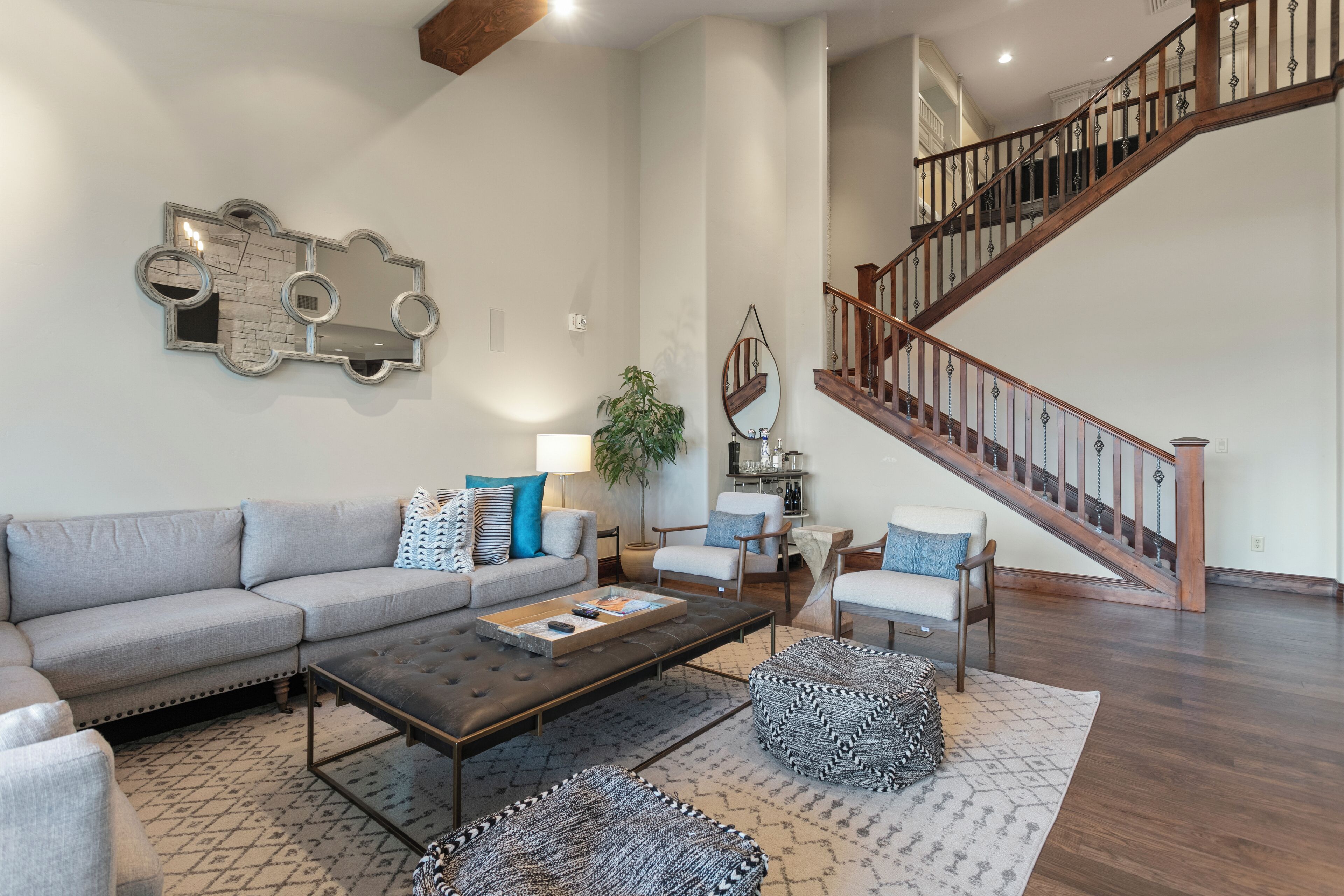 A spacious living room with a gray sectional sofa, a coffee table, and patterned ottomans. A wooden staircase with a railing is visible in the background.