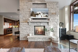 A modern living room with a stone fireplace, a flat-screen TV mounted above it, and a view of a snowy landscape. - Ski-in/ski-out At Deer Valley 3br 3ba 3 Bedroom Condo (Park City)