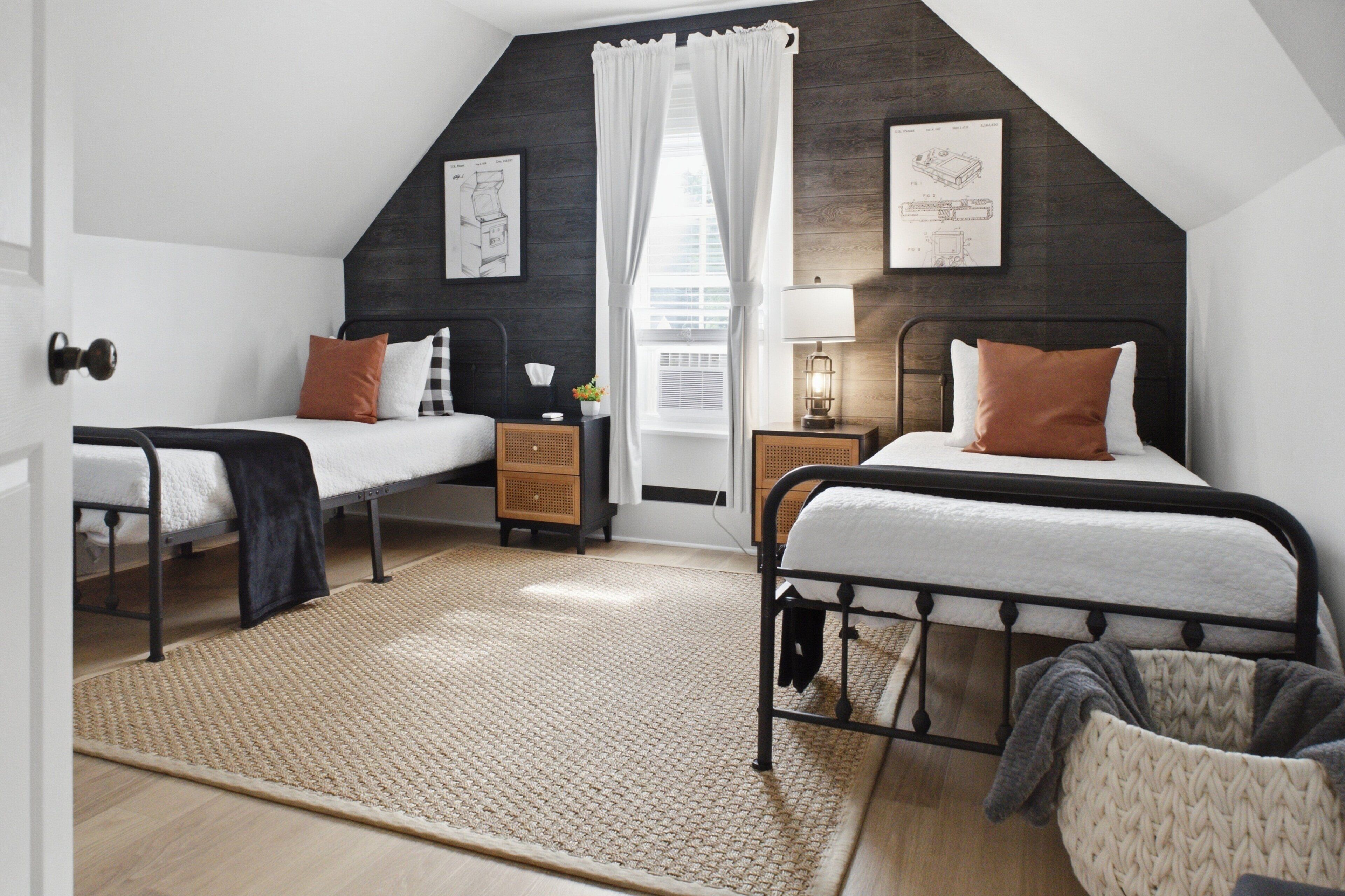 A bedroom with two beds, a window with curtains, a bedside table with a lamp, and a wall-mounted artwork.