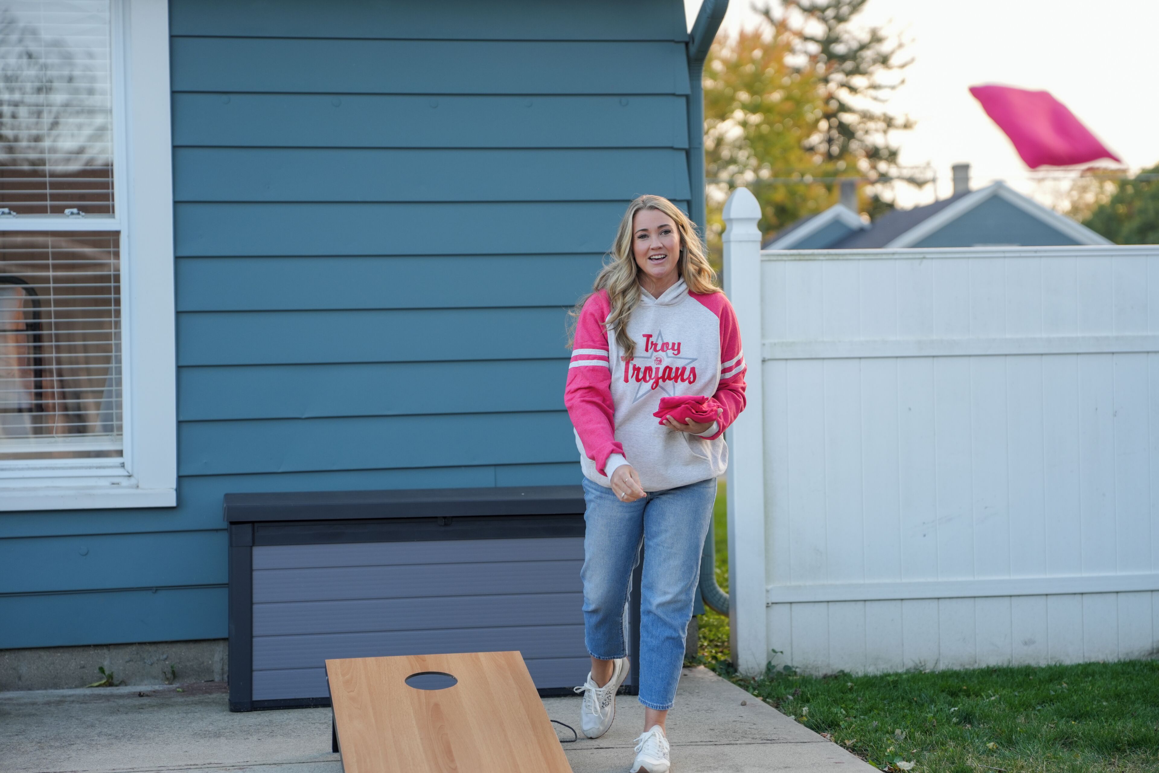 A person standing next to a wooden board, wearing a jacket with 'Troy Trojans' on it, in front of a house with blue siding and a white fence.