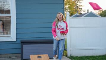 A person standing next to a wooden board, wearing a jacket with 'Troy Trojans' on it, in front of a house with blue siding and a white fence.