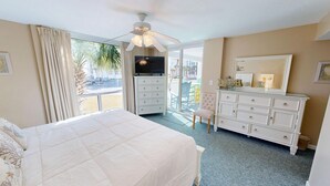Habitación de hotel con cama, cómoda, televisor y balcón con vista a palmeras y un estacionamiento.