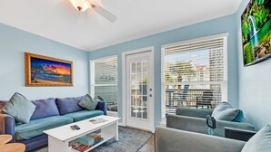 Ein Wohnzimmer mit einem blauen Sofa, einem weißen Couchtisch und einem grauen Schlafsofa. Zwei Fenster mit Jalousien, ein Gemälde an der Wand und ein Deckenventilator sind vorhanden.