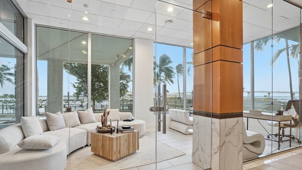 A modern living room with a curved sofa, a wooden coffee table, and a glass wall offering a view of palm trees and a beach. - Luxurious Waterfront Condo At Alta Mar 2 Bedroom Condo (Fort Myers)