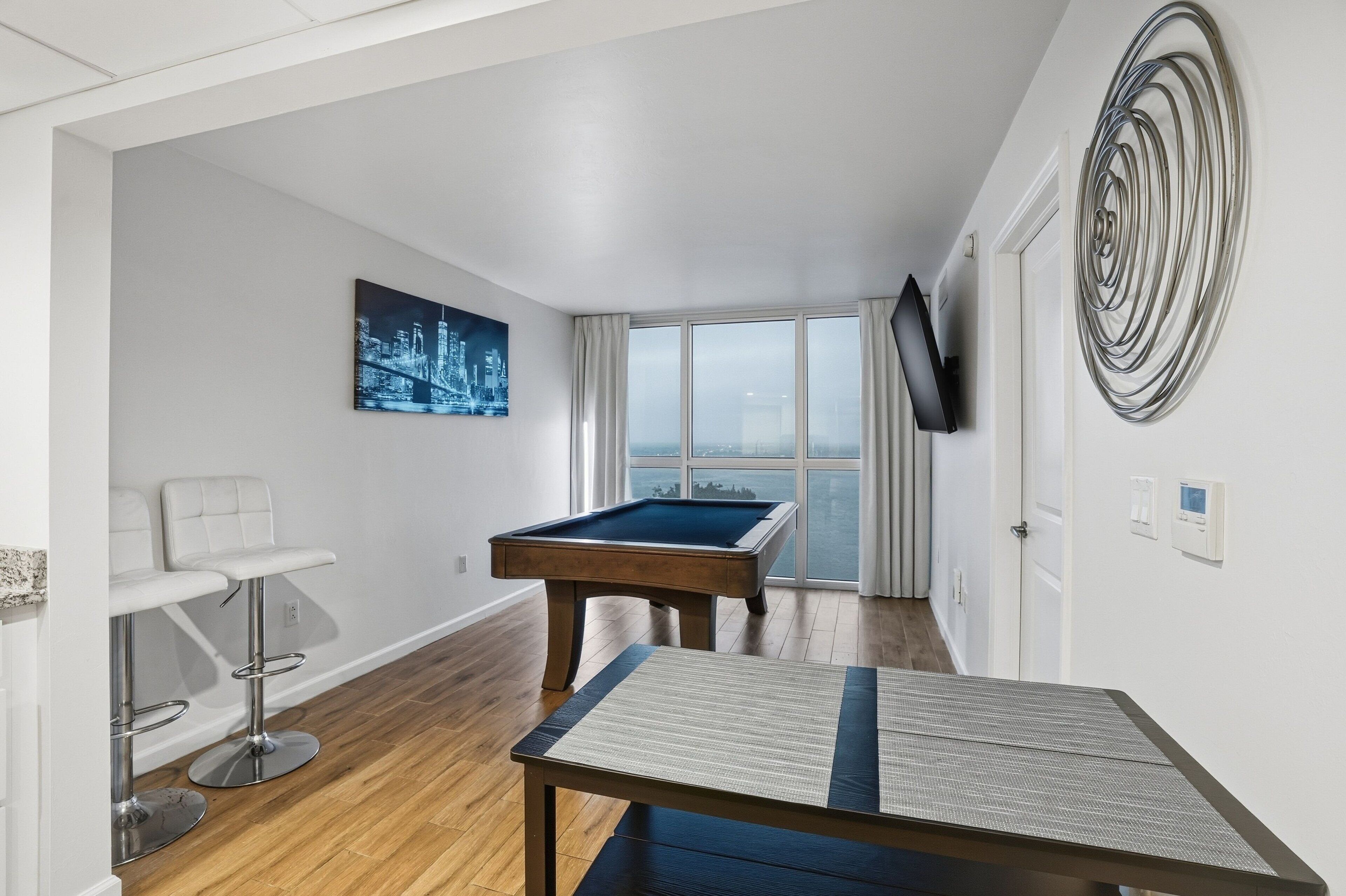 Ein modernes Wohnzimmer mit Billardtisch, Bar mit Barhocker, großem Fenster und Fernseher an der Wand.