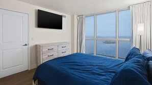 A bedroom with a large window, a bed with blue bedding, a white dresser, and a flat-screen TV mounted on the wall. - Luxurious Waterfront Condo At Alta Mar 2 Bedroom Condo (Fort Myers)