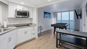 A modern kitchen with white cabinets, a stainless steel microwave, a granite countertop, and a pool table in a room with large windows and a cityscape view. - Luxurious Waterfront Condo At Alta Mar 2 Bedroom Condo (Fort Myers)