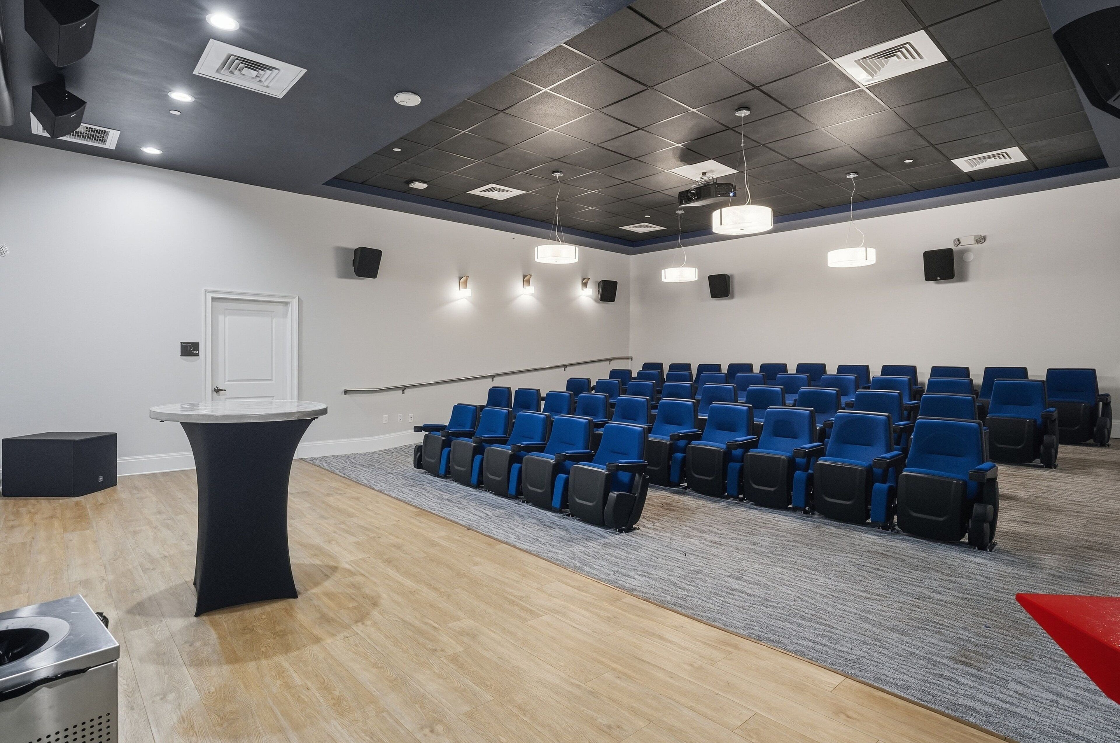 Una sala conferenze con file di sedie blu, una porta bianca, un tavolo nero e pavimento rivestito di moquette.
