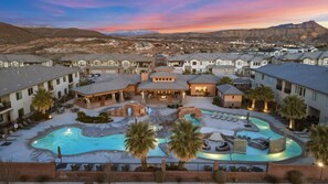 Un resort con varias edificaciones, un área de piscina central con jacuzzi y vista a las montañas al fondo.