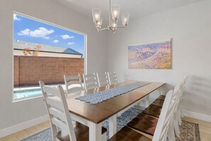 Comedor con mesa de madera, sillas blancas, candelabro, vista de un edificio a través de una ventana y un cuadro en la pared.