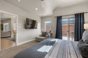 Un dormitorio con una cama grande, un televisor montado en la pared, una puerta corrediza de vidrio al exterior y un pequeño rincón de descanso con una silla y un otoman.