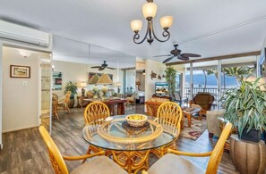 A spacious living area with a round glass table, wicker chairs, a ceiling fan, and a view of the ocean. - Menehune Shores, #308 2 Bedroom Condo (Kihei)