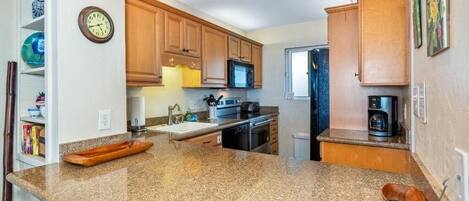A kitchen with granite countertops, wooden cabinets, a microwave, a coffee maker, and a clock on the wall.